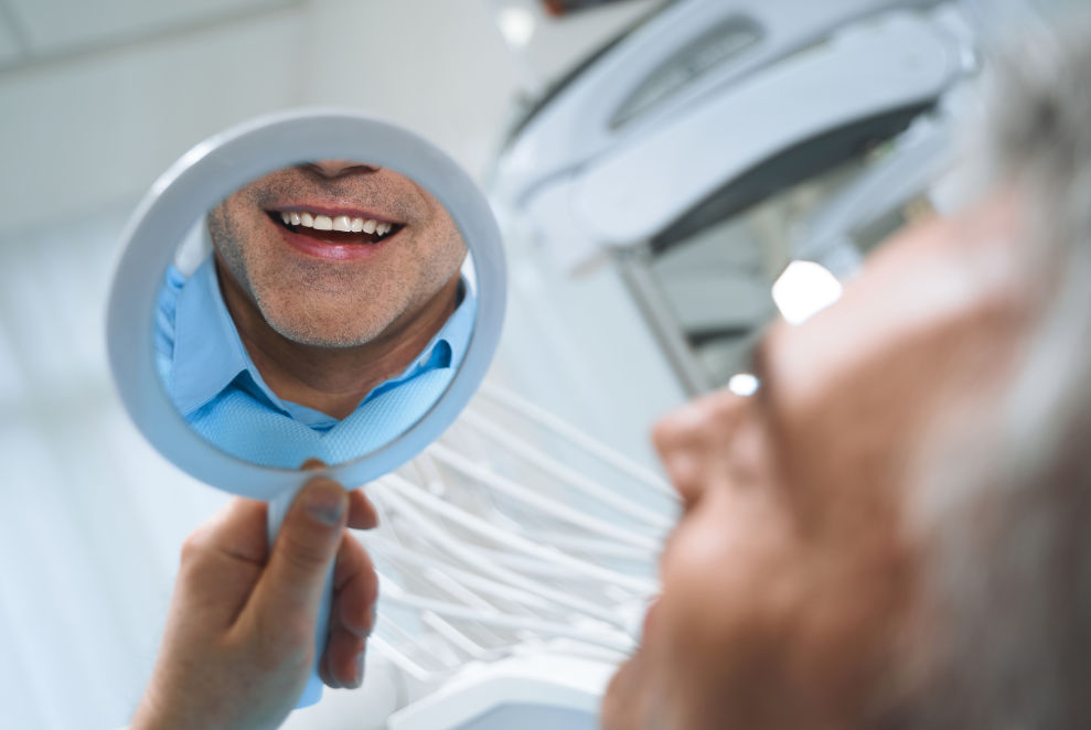 male patient with dental implants smiling in a hand mirror at the dental office